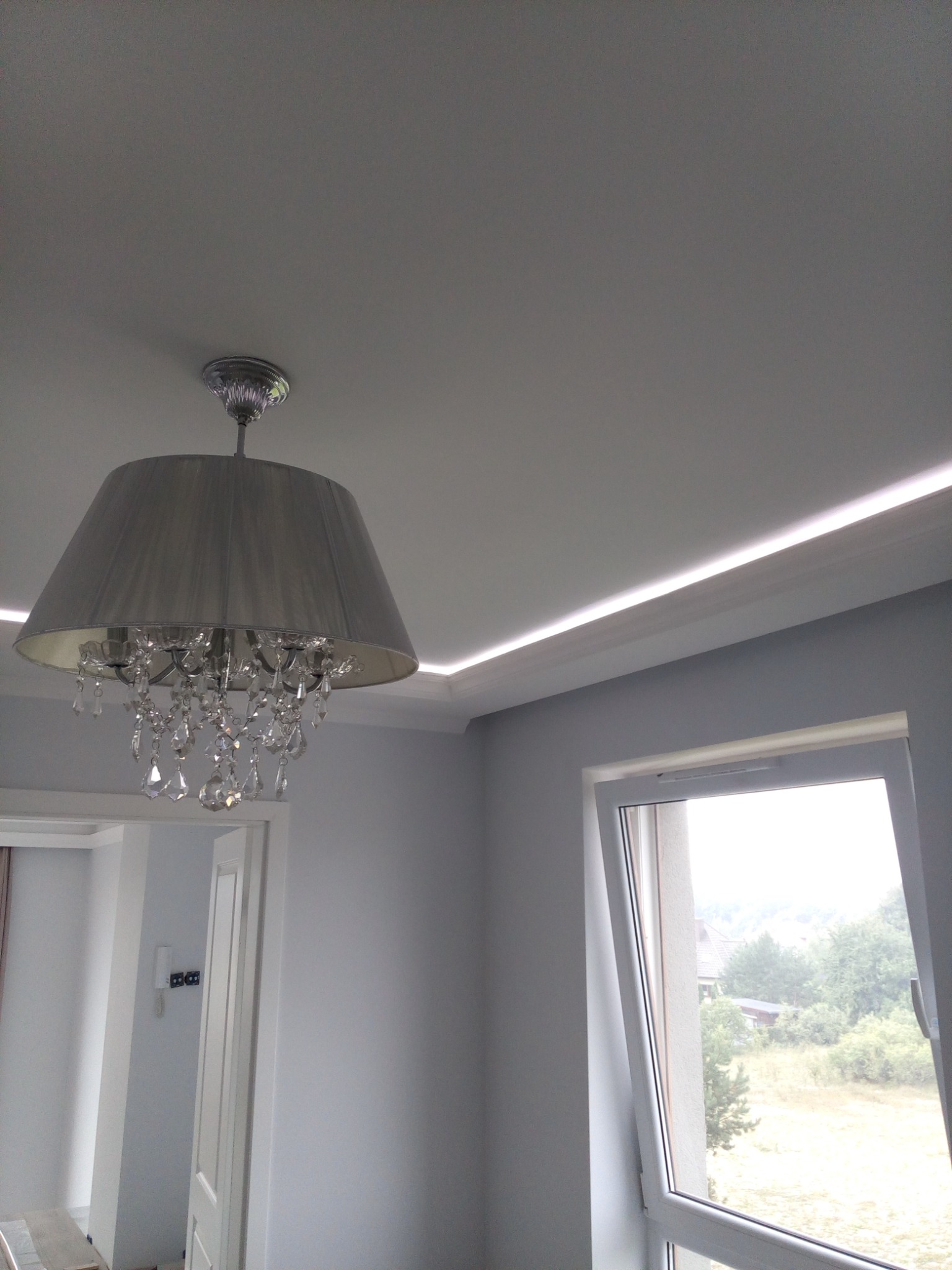 Szaro-białe wnętrze z kryształowym żyrandolem w abażurze i podświetleniem LED wzdłuż sufitu, widok na okno z zielenią na zewnątrz.