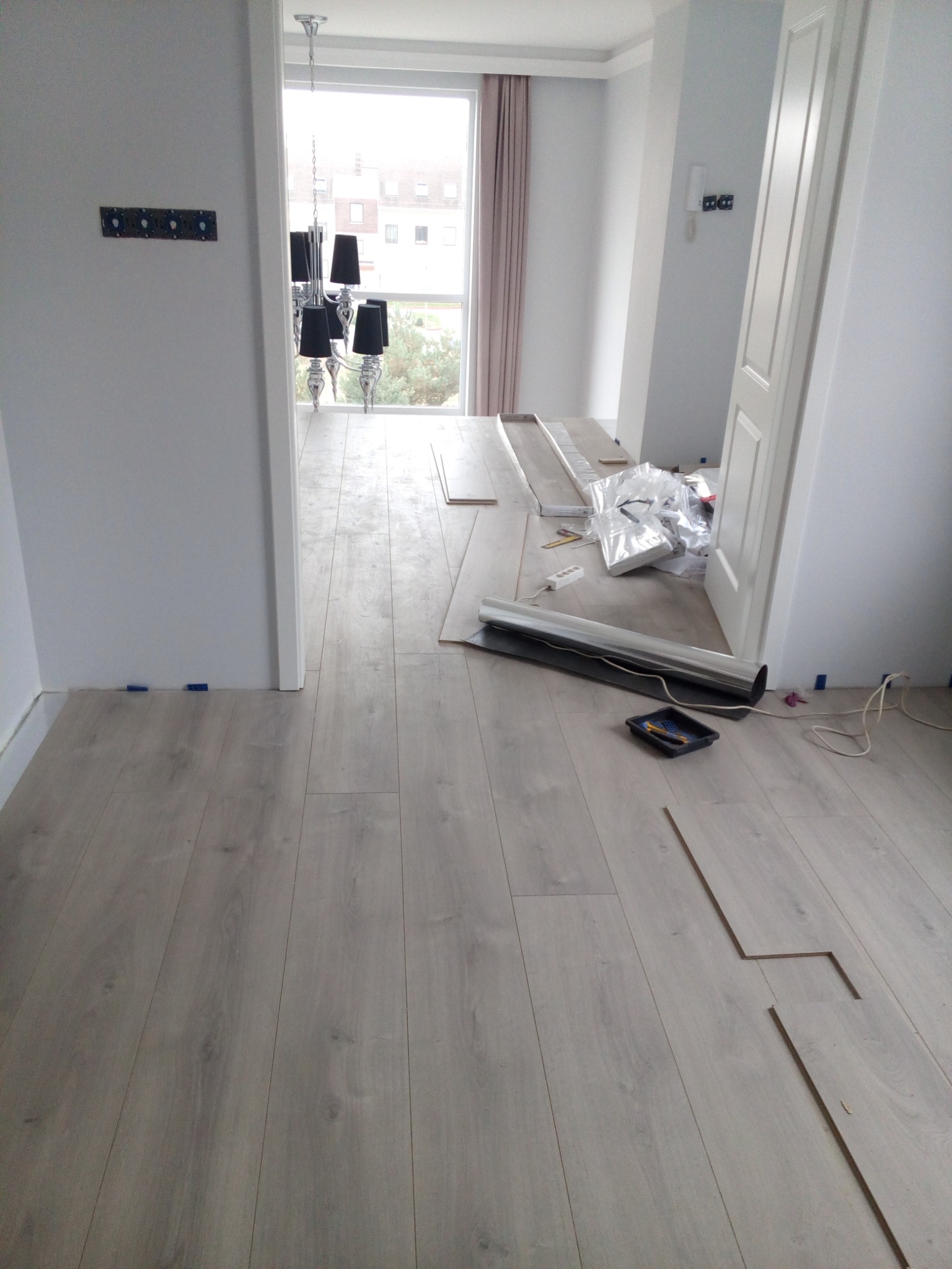 Układanie paneli podłogowych w jasnym pomieszczeniu, widoczne narzędzia i materiały przygotowane do montażu, w tle okno z widokiem na budynki.