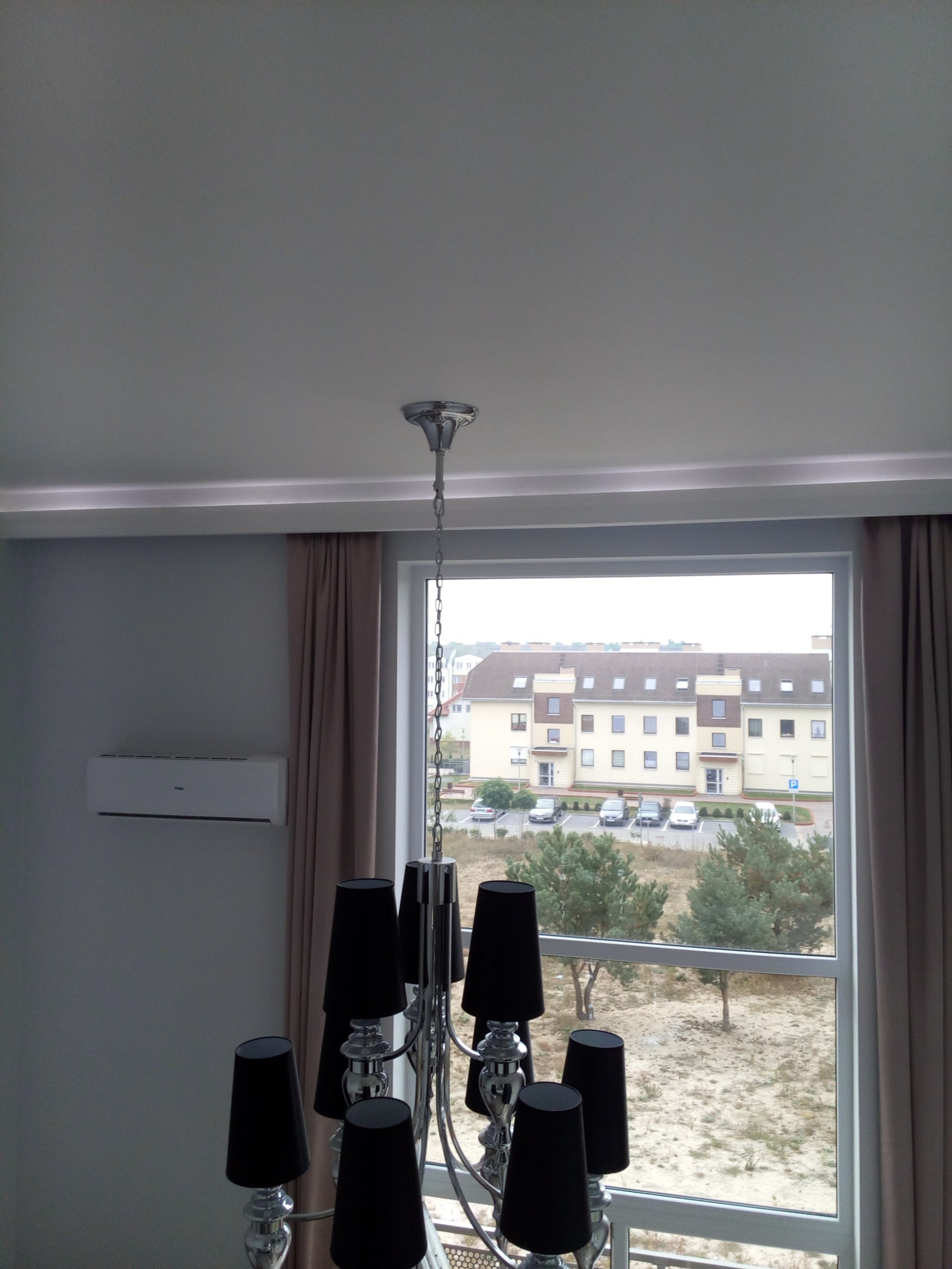 Nowoczesny salon z klimatyzatorem i srebrnym żyrandolem z czarnymi abażurami, widok z okna na parking i budynek mieszkalny.