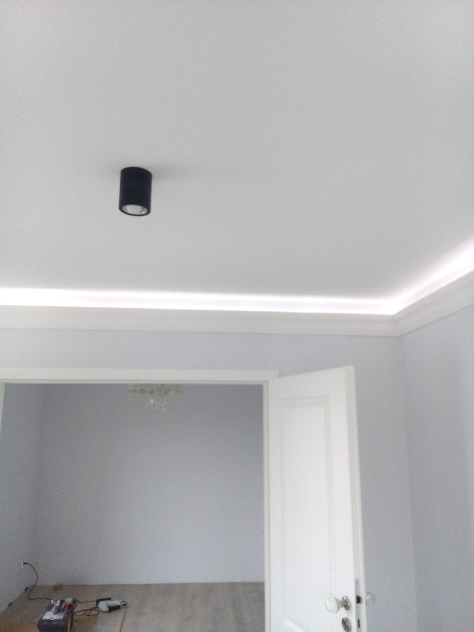 Białe wnętrze z listwami LED przy suficie, czarnym reflektorem punktowym i otwartymi drzwiami do pokoju z drewnianą podłogą i żyrandolem, widoczne narzędzia na podłodze sugerują prace wykończeniowe.