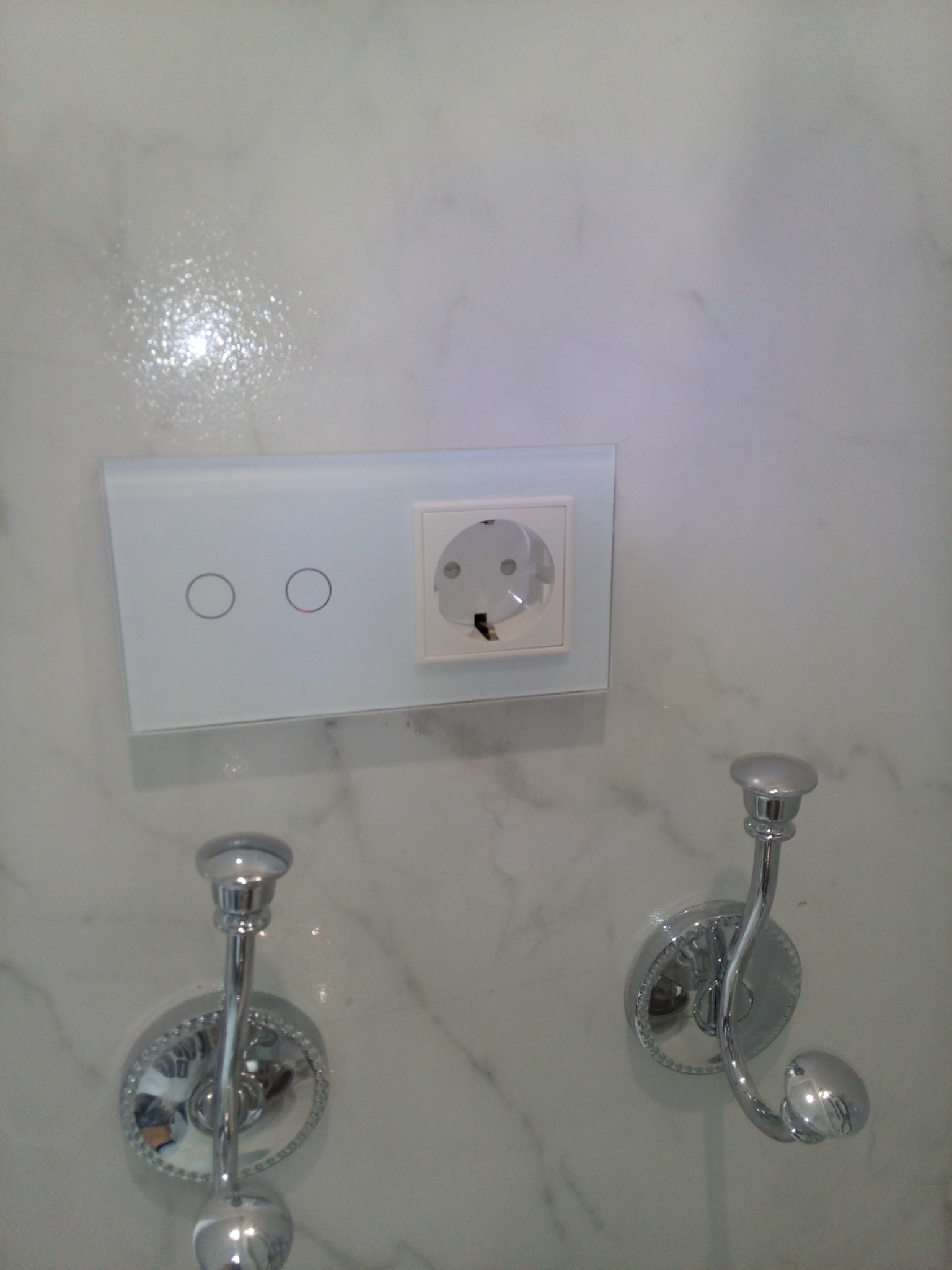 Biała, marmurowa ściana z zamontowanym szklanym panelem dotykowym z dwoma okrągłymi włącznikami światła i gniazdkiem elektrycznym oraz dwoma chromowanymi wieszakami na ręczniki o dekoracyjnych...