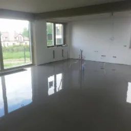 Świeżo wylana, gładka, szara posadzka w przestronnym pomieszczeniu z dużymi oknami, odbijającymi krajobraz zewnętrzny i niebo. Widoczne wystające rurki instalacyjne.