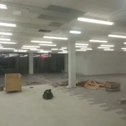 Przestrzeń komercyjna w trakcie prac wykończeniowych posadzki; widoczna część wylewki oraz fragmenty ułożonej szarej nawierzchni z wzorem, kartony, narzędzia i torba robocza.