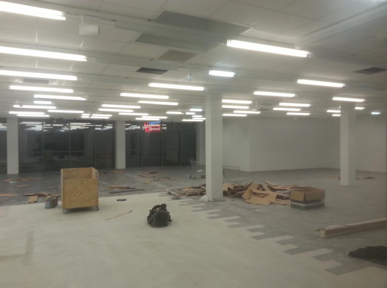 Przestrzeń komercyjna w trakcie prac wykończeniowych posadzki; widoczna część wylewki oraz fragmenty ułożonej szarej nawierzchni z wzorem, kartony, narzędzia i torba robocza.