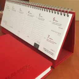 Czerwony kalendarz biurkowy ze spiralą i czerwony notatnik z wytłoczonym logo firmy MEAT-PROS na drewnianym biurku, widok z bliska.