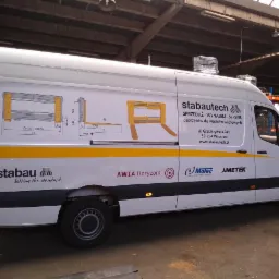 Biały samochód dostawczy z grafiką przedstawiającą schematy osprzętu do wózków widłowych oraz logo firm Stabautech, Stabau, Awia Horyzont, Molec i Ametek.