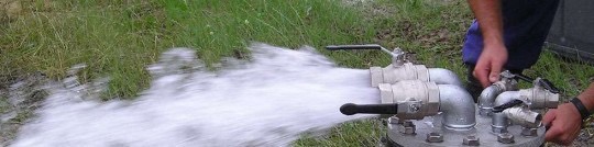 Wypływająca woda z metalowej instalacji z zaworami, trawa w tle.