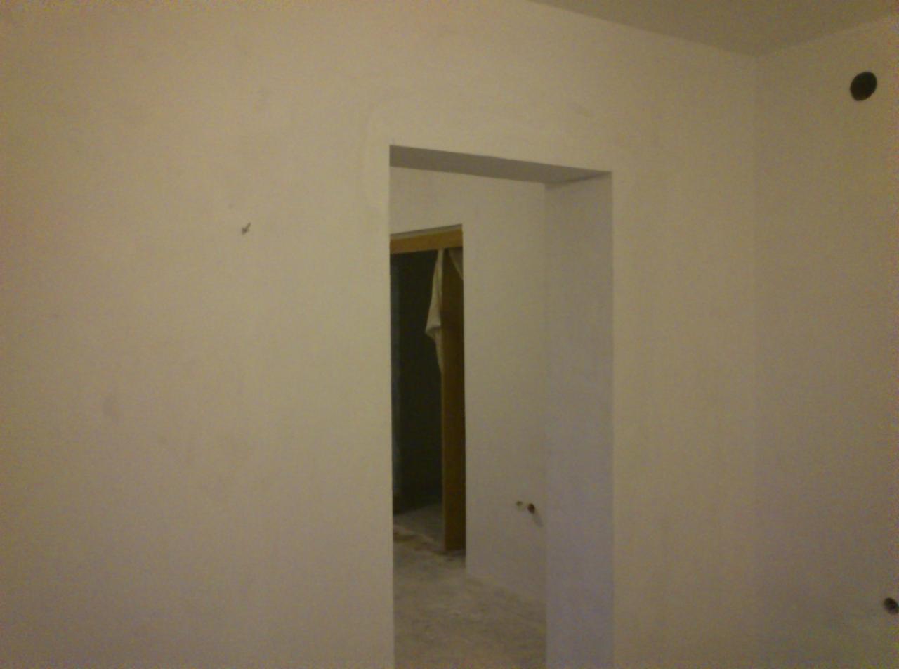 Gładka, świeżo pomalowana ściana w odcieniach bieli z widocznym przejściem do innego pomieszczenia przez futrynę drzwiową. Widoczne ślady po gniazdkach.