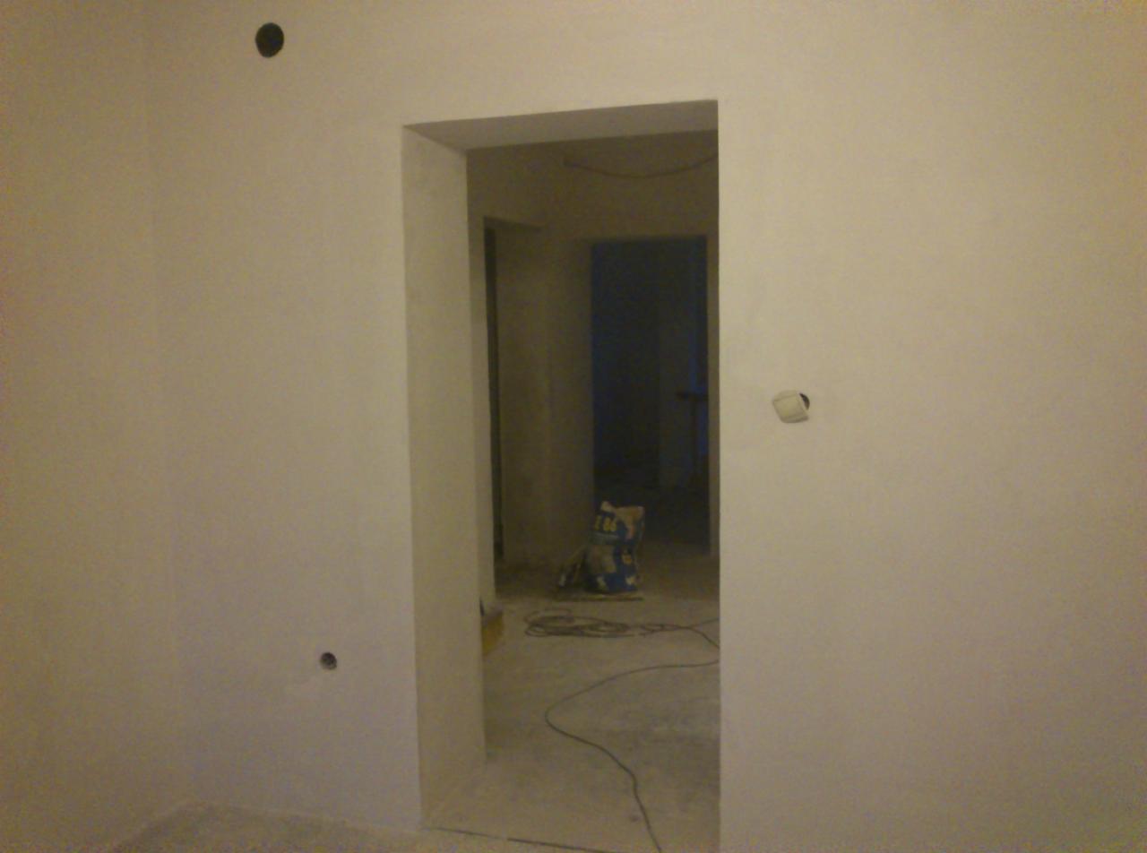 Wnętrze po gładziowaniu: gładkie, białe ściany z widocznymi puszkami elektrycznymi i otworem drzwiowym. Na podłodze kabel i worek zaprawy.