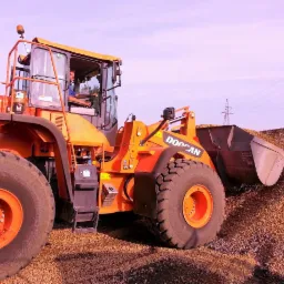 Pomarańczowa ładowarka kołowa marki Doosan z pełną łyżką żwiru na tle błękitnego nieba z widocznymi liniami wysokiego napięcia.