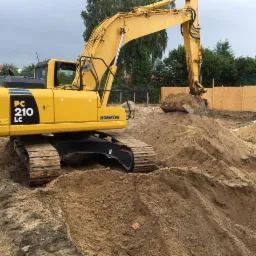 Żółta koparka Komatsu PC210 LC podczas prac ziemnych, wykop pod fundamenty, widoczna pryzma piasku i częściowo drewniane szalunki.