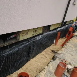 Prace związane z izolacją fundamentów budynku: widoczna czarna folia kubełkowa, rury drenarskie, piasek zasypowy, oraz pracownicy z ładowarką.