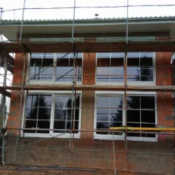 Dom w trakcie budowy z zamontowanymi oknami, otoczony rusztowaniem, widoczne samochody i drzewa w tle.