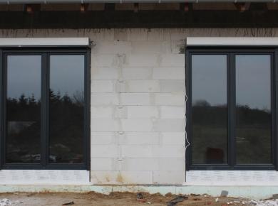 Dwa czarne okna z roletami zewnętrznymi w surowym murze z betonu komórkowego, widok z zewnątrz, jesienne tło za szybami.