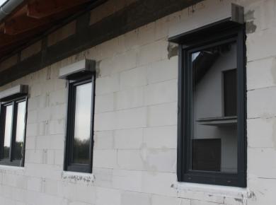 Czarne okna z roletami zewnętrznymi w bloku z betonu komórkowego. Widoczne trzy okna, z czego jedno jest otwarte.