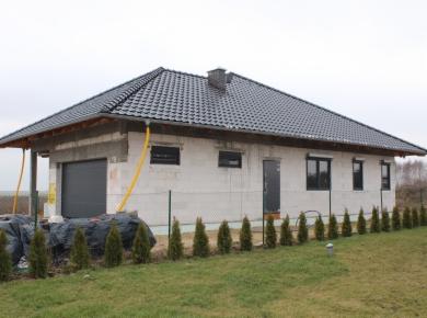 Dom jednorodzinny w stanie surowym zamkniętym z czarną dachówką, widoczne okna z ciemnymi ramami i brama garażowa, otoczony młodymi tujami.