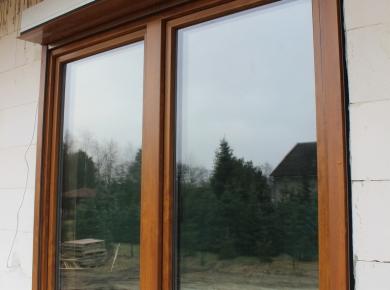 Dwuokienne okno z brązową ramą i roletą zewnętrzną, odbijające w szybach drzewa i budynek. Widok z zewnątrz na ścianę z białych pustaków.