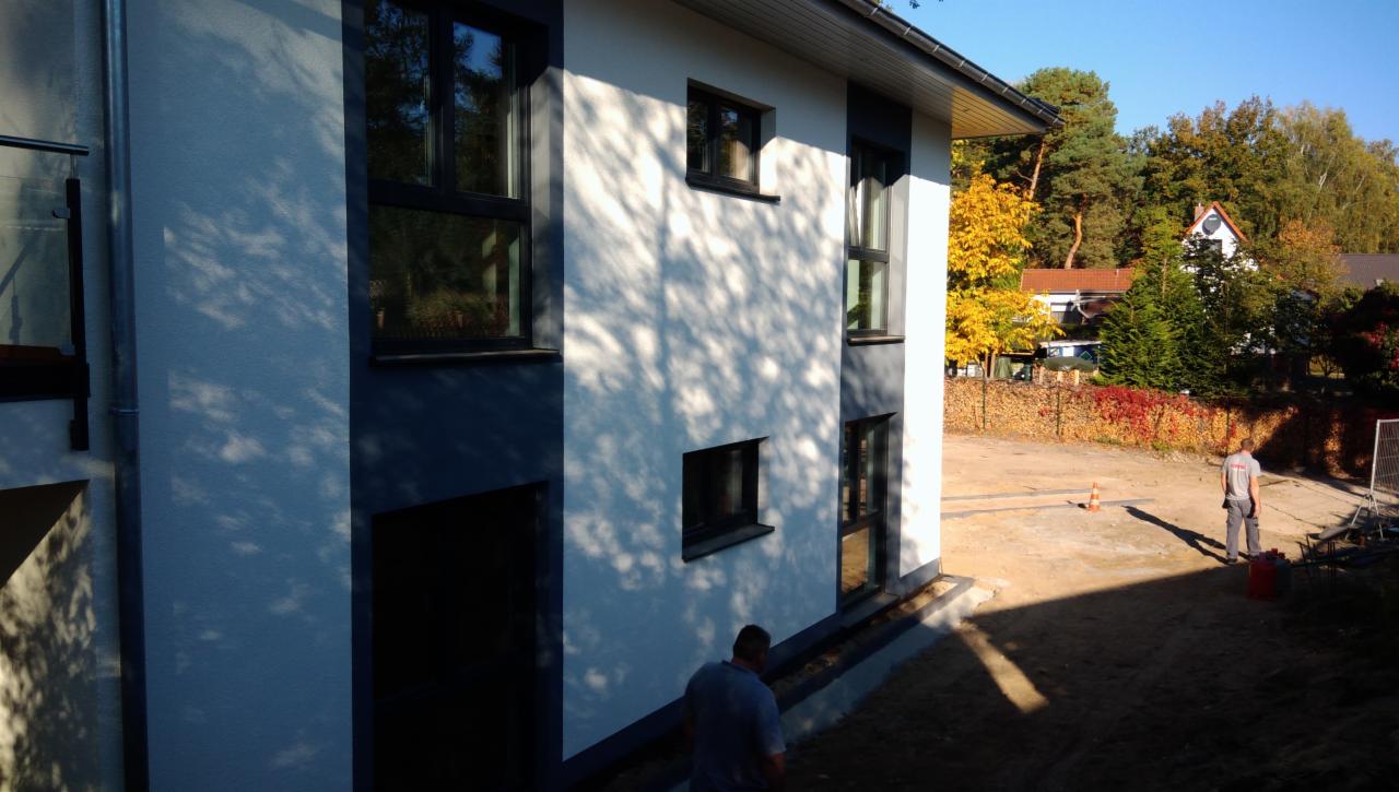 Nowoczesny budynek z białymi ścianami i ciemnymi oknami w trakcie wykończenia, widoczne cienie drzew na elewacji i robotnicy na zewnątrz.