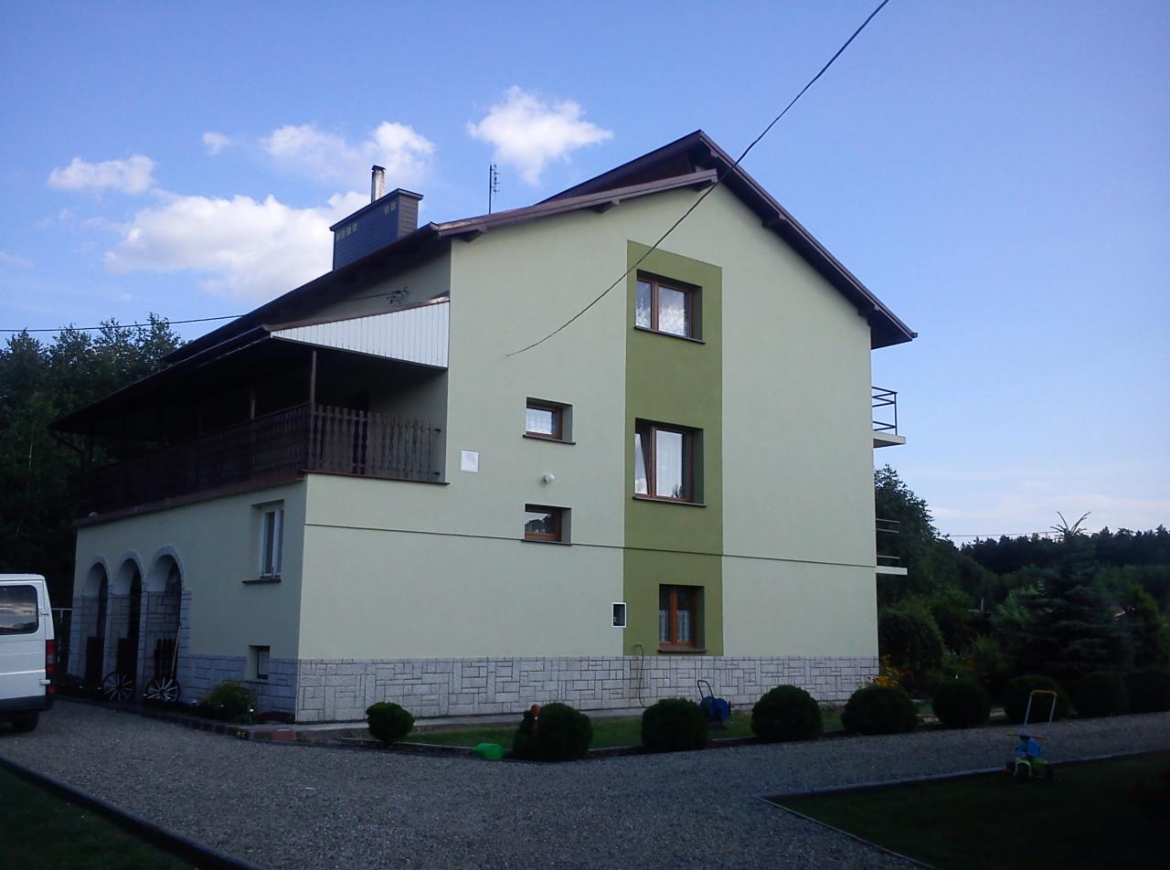 Dwukondygnacyjny dom z elewacją w odcieniach zieleni, widoczny z perspektywy ogrodu, z elementami dekoracyjnymi z kamienia i drewnianym balkonem.