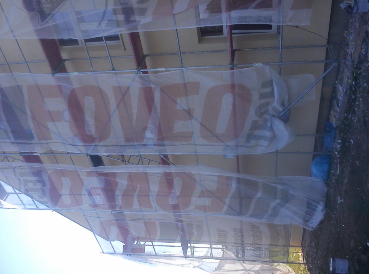 Elewacja budynku w trakcie prac, osłonięta siatką z reklamą firmy. Widoczne rusztowania i fragment okna. Ujęcie z dołu, pod kątem.