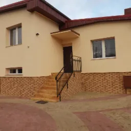 Elewacja domu jednorodzinnego w beżowym kolorze z ceglaną podmurówką i schodami wejściowymi z czarną balustradą. Widoczny fragment chodnika z kostki.