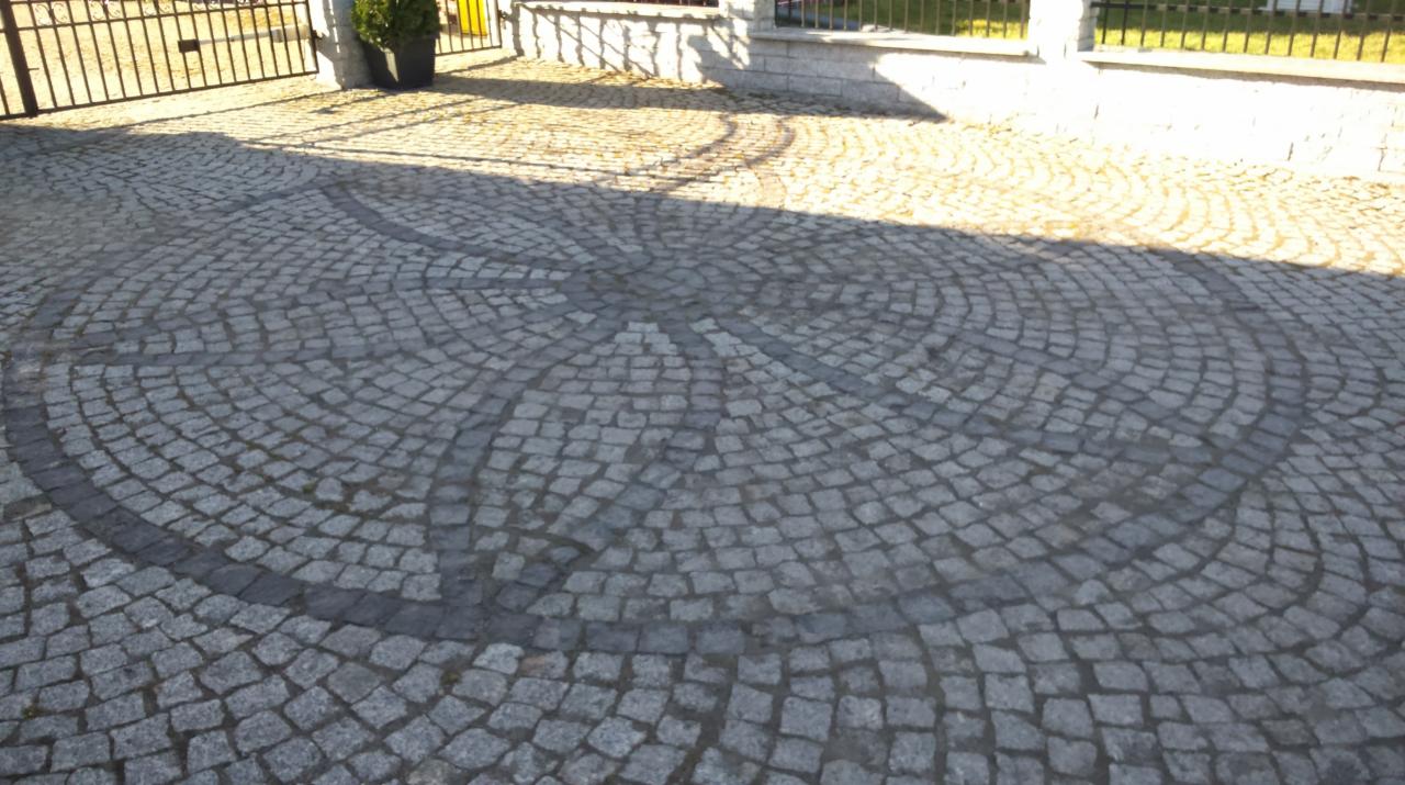 Ułożona kostka brukowa na podjeździe z dekoracyjnym wzorem koła, częściowo zacieniona, z widocznym fragmentem ogrodzenia i rośliny w tle.