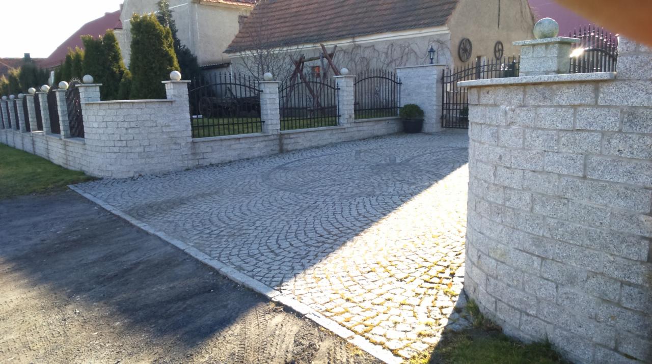 Granitowa kostka brukowa ułożona w łukowy wzór, ogrodzenie z kamiennymi słupkami i kutą bramą, zieleń w tle, słoneczny dzień.