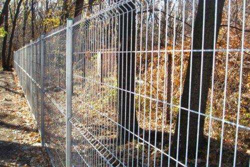 Długi, metalowy płot z siatki zgrzewanej, z zielonymi zaślepkami na słupkach, oddzielający ścieżkę od zarośli i drzew w jesiennej scenerii.