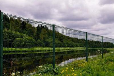 Zielone, metalowe ogrodzenie panelowe z widokiem na staw i las w tle, pochmurne niebo.