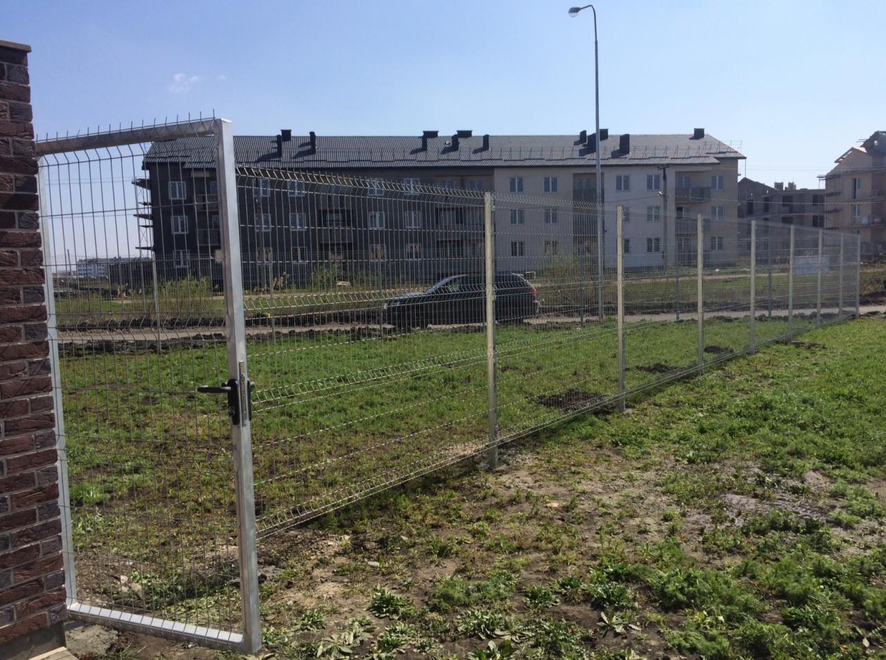 Metalowa siatka ogrodzeniowa z furtką, widoczna na tle budynku mieszkalnego i trawnika z ubytkami darni.