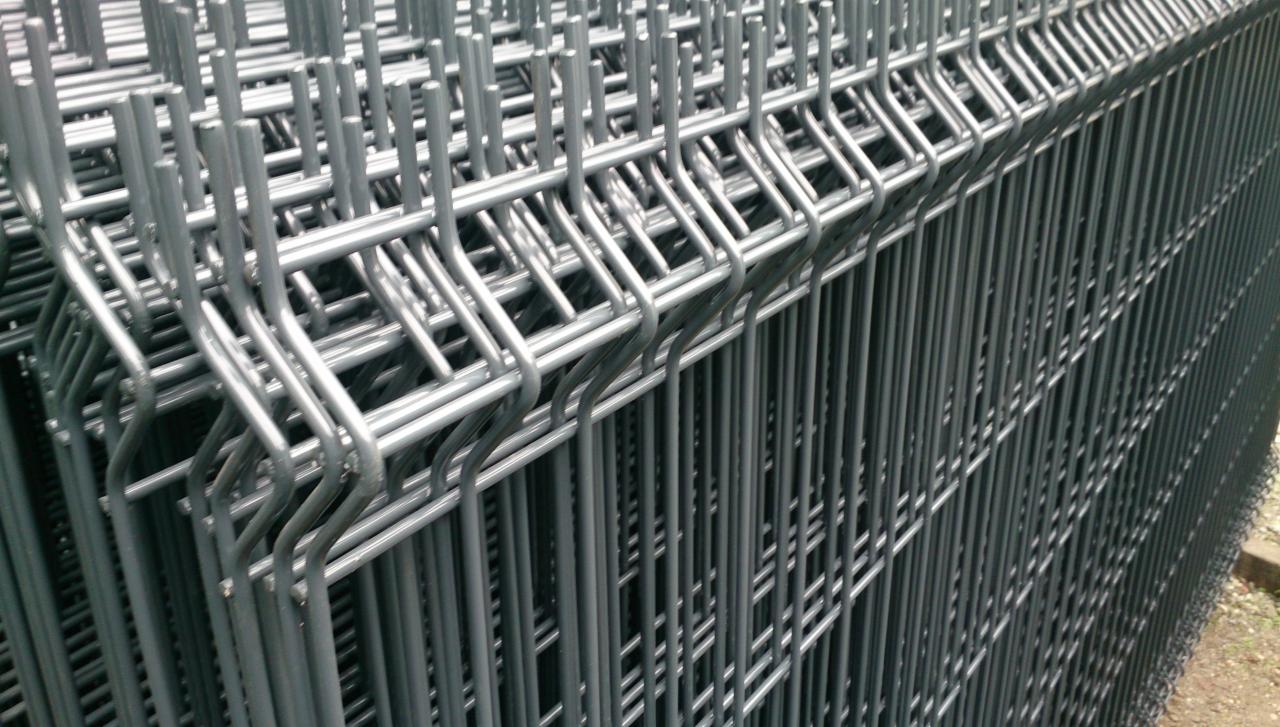 Stos szarych, spawanych paneli ogrodzeniowych z drutu, widocznych pod kątem, z zielonym tłem w oddali.