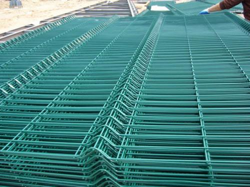 Stos zielonych, spawanych paneli ogrodzeniowych z drutu, układanych jeden na drugim, widoczna ręka w niebieskiej rękawicy układająca panele.