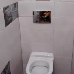 Biała wisząca toaleta z chromowanym przyciskiem spłukującym na tle jasnych płytek. Dekoracyjne płytki z motywem retro po lewej stronie. Widoczny fragment podłogi.