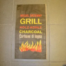 Brązowa papierowa torba z nadrukiem: 'Węgiel Drzewny Grill Holz-Kohle Charcoal Carbone di Legna' i motywem płomieni na tle szarej ściany z płytek.