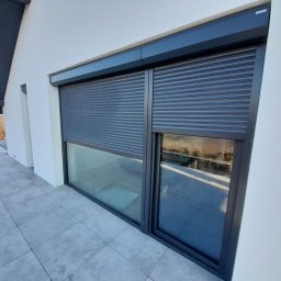 PATRYK FRĄCZ 7Trade HOUSE - Zewnętrzna roleta antywłamaniowa w kolorze antracytowym, częściowo opuszczona, zamontowana na oknie nowoczesnego domu z widokiem na krajobraz.