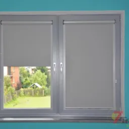 Szare, zwinięte plisy w oknie z widokiem na ogród i budynek w tle, na turkusowej ścianie, z widocznym logo firmy w prawym dolnym rogu.