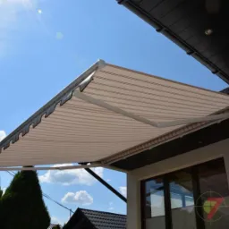 Beżowa markiza z delikatnymi paskami zamontowana nad oknem domu w Olszowicach, widoczna z dołu pod kątem, błękitne niebo w tle.