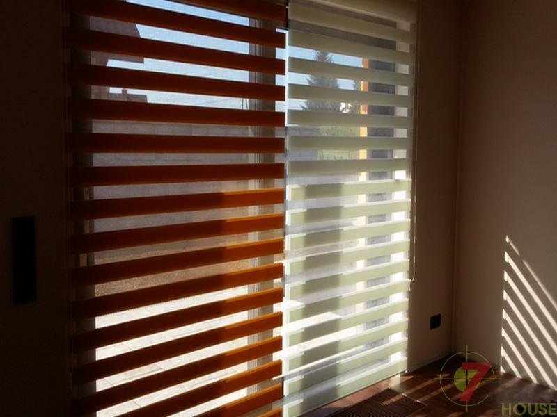Dwuczęściowe okno z zamontowanymi roletami typu dzień i noc, jedna część w odcieniach brązu, druga w jasnej tonacji, z widocznymi promieniami słonecznymi wpadającymi do wnętrza.