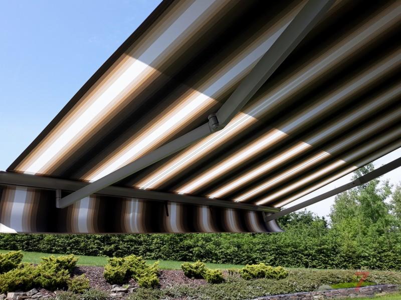 Rozwinięta markiza w paski (brąz, beż, biel) zamontowana nad tarasem, widok od dołu, na tle zieleni i błękitnego nieba.