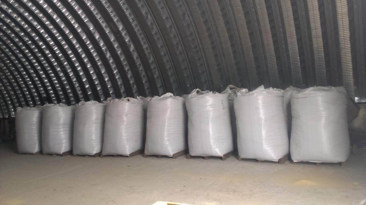 Rząd białych, worków z pelletem na paletach w magazynie o metalowej konstrukcji łukowej. Widok z przodu, oświetlenie magazynowe.