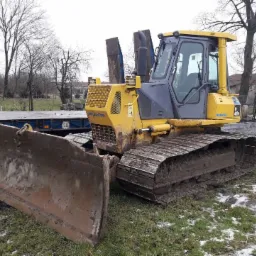 Żółty buldożer Komatsu D41P, częściowo zabłocony, stoi na trawiastym terenie w Sławnie, z widoczną naczepą transportową w tle i budynkiem mieszkalnym w oddali.