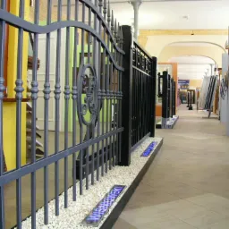 Wystawa elementów ogrodzeniowych: ozdobne, metalowe przęsła na białym żwirze, podświetlane logo producenta, ekspozycja w hali z oknami i drzwiami.