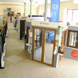 Ekspozycja okien i drzwi w salonie sprzedaży z kolumnami. Widoczne różne modele i kolory, logo Vetrex i Veka. Duże okno z prawej strony.