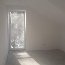 MK Firma Usługowa Michał Kohnke - Pusty pokój na poddaszu z oknem i świeżo wylana wylewką podłogową, oświetlony silnym światłem słonecznym, widoczny kabel wystający z sufitu.