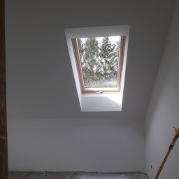 MK Firma Usługowa Michał Kohnke - Poddasze po malowaniu na biało, widoczne okno dachowe z widokiem na drzewa, częściowo zakryta podłoga i narzędzie malarskie