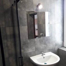 MK Firma Usługowa Michał Kohnke - Szara łazienka z kabiną prysznicową z czarnymi profilami, białą umywalką i lustrem, w którym odbija się fragment kuchni.