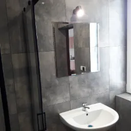 Szara łazienka z kabiną prysznicową z czarnymi profilami, białą umywalką i lustrem, w którym odbija się fragment kuchni.