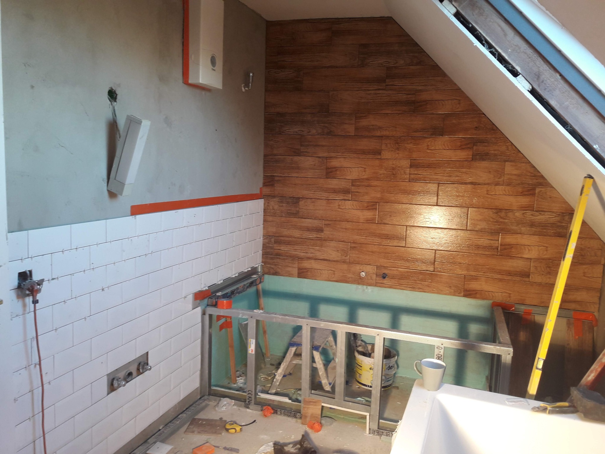 Wnętrze łazienki w trakcie remontu, z widocznymi płytkami imitującymi drewno na ścianie pod skosem, białe płytki na dolnej części ściany, metalowa konstrukcja pod zabudowę wanny oraz poziomica...