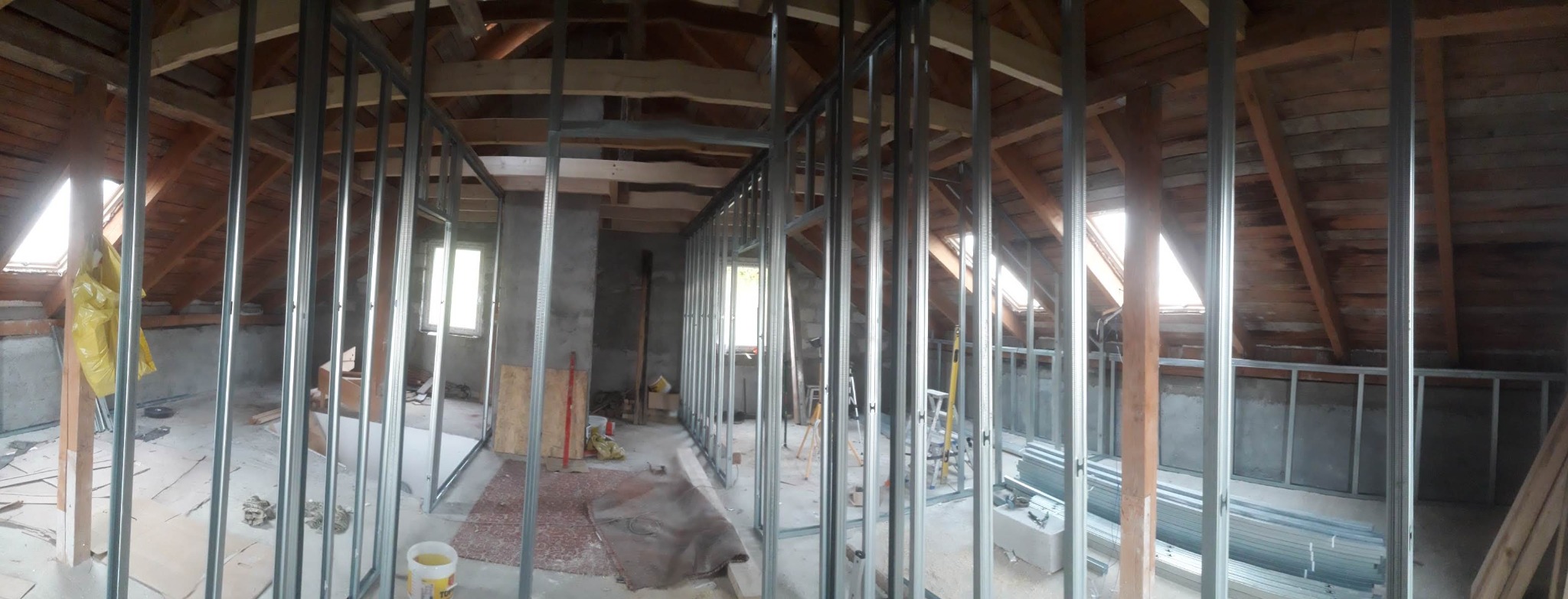 Szkielet ścian działowych z profili metalowych w surowym stanie poddasza z oknami dachowymi, widoczne drewniane elementy konstrukcyjne dachu i przygotowane materiały budowlane.