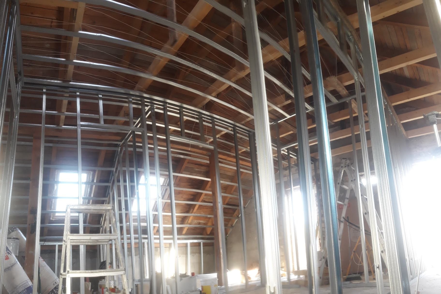 Metalowa konstrukcja szkieletowa pod zabudowę z płyty karton-gips na poddaszu, widoczne drewniane elementy więźby dachowej i okno dachowe, na podłodze częściowo widoczne materiały budowlane.